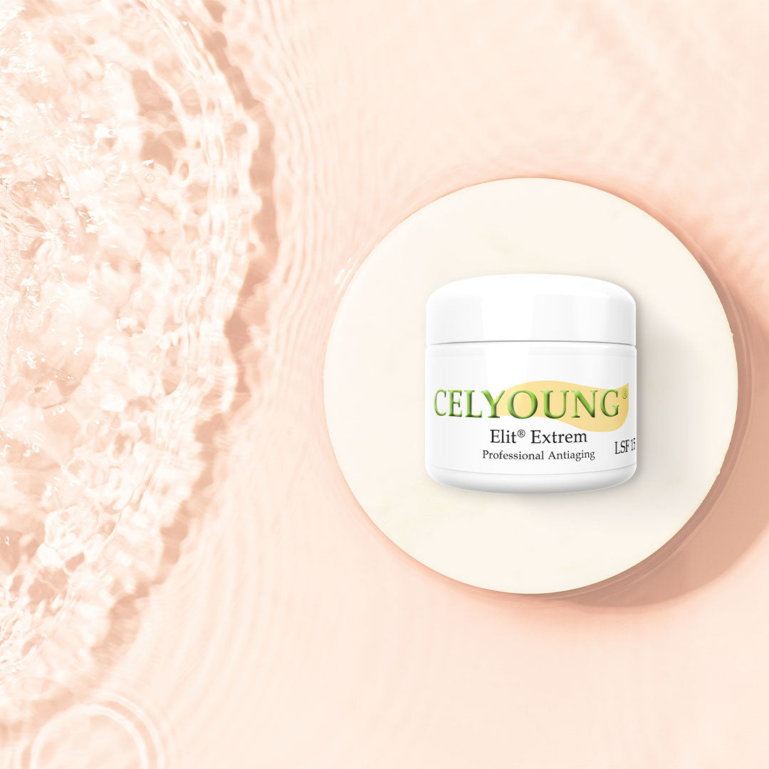 Ein weißer Tiegel mit Celyoung Elit Extrem Creme LSF 15 steht auf einer runden, hellen Plattform mit sanft plätscherndem Wasser auf der linken Seite und einem hellpfirsichfarbenen Hintergrund und präsentiert dieses professionelle Anti-Aging-Hautpflegeprodukt.